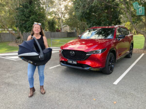 A woman standing next to a red suv with a baby carrier.