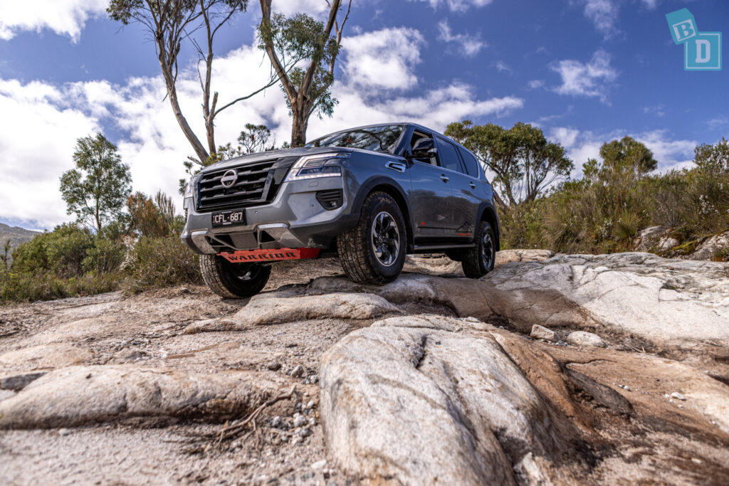The 2023 Nissan Patrol Warrior 4x4 is driving on rocks in the forest.