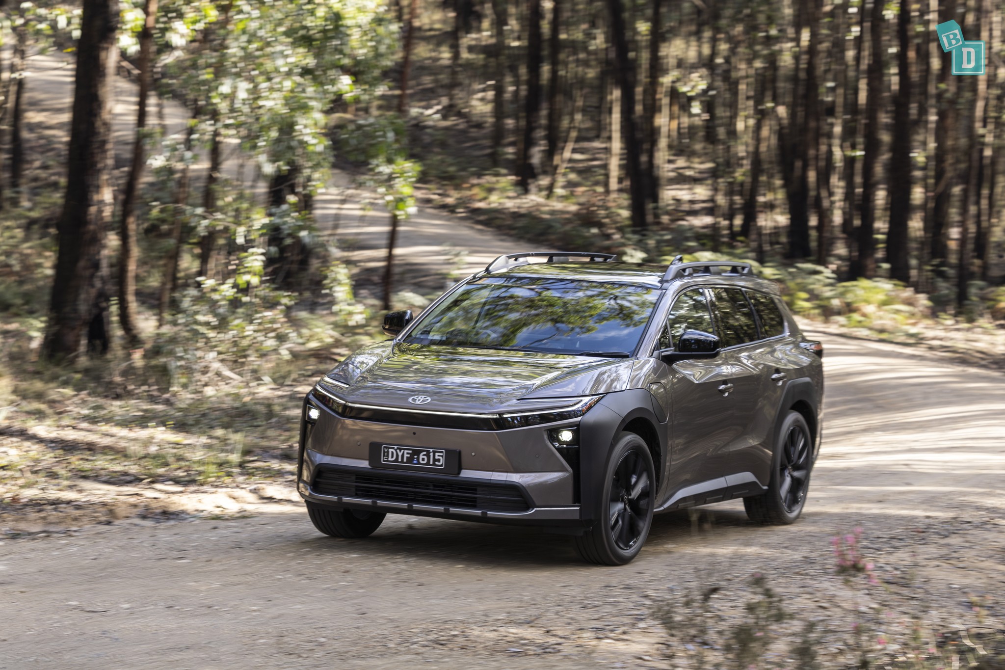 2026 Toyota bZ4X Touring front three-quarter view driving on a dirt road through a sunlit forest